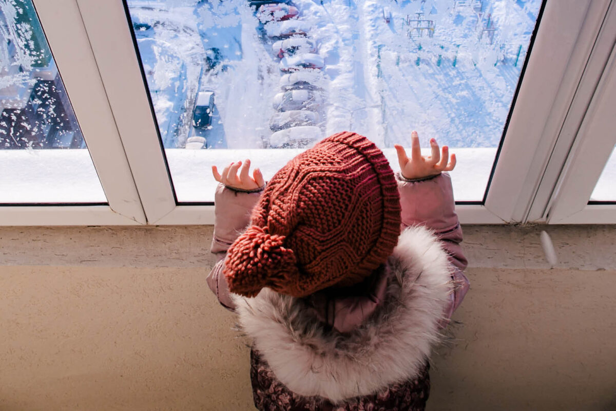 Kind schaut aus dem Fenster auf eine verschneite Straße und Landschaft