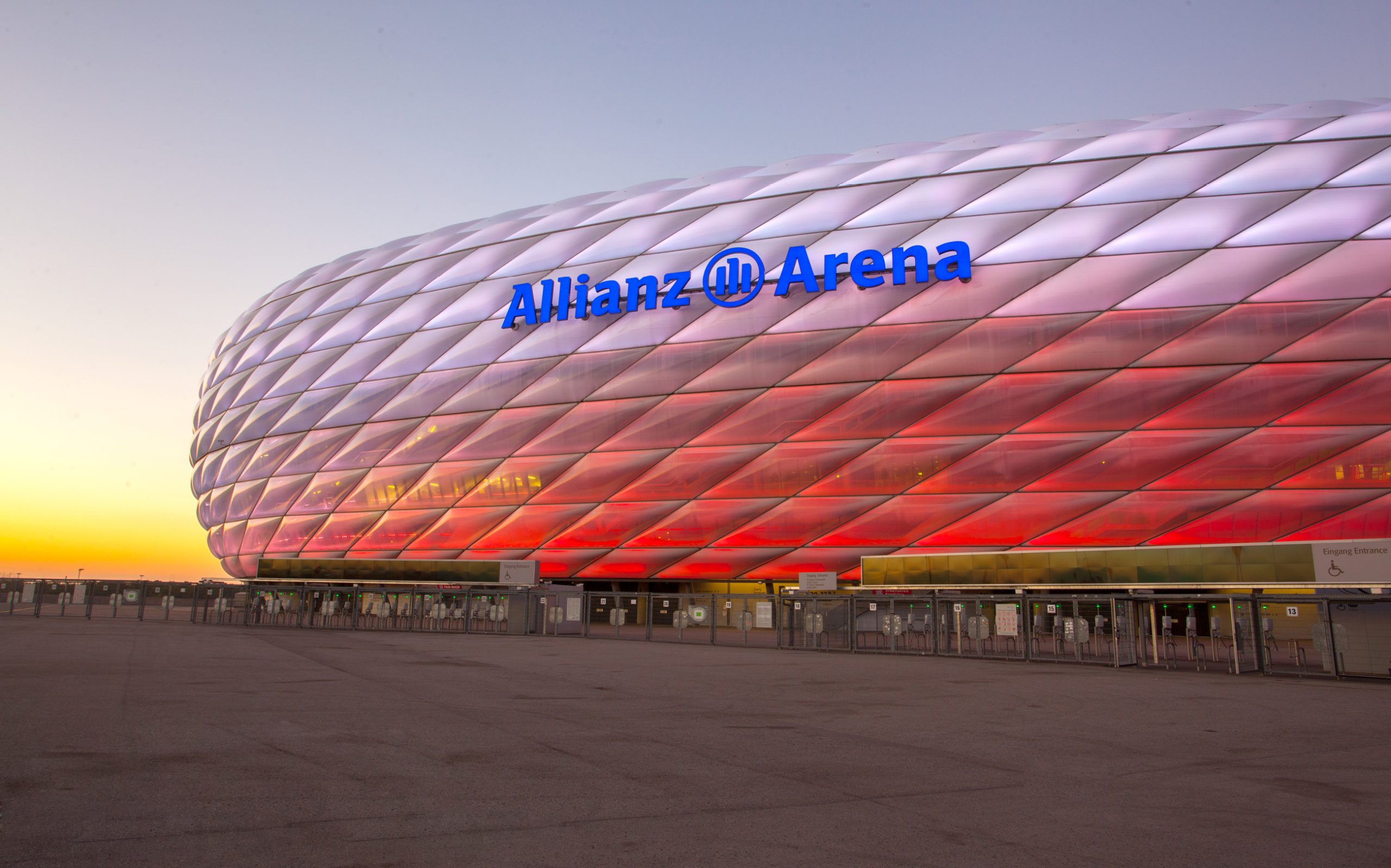 Außenansicht der Münchner Allianz Arena in der Abenddämmerung: Die charakteristische Kissenfassade des Stadions leuchtet im unteren Bereich in Rot und geht nach oben in Weiß über. Im Vordergrund erstreckt sich der weitläufige, leere Vorplatz mit den Einlassbereichen und Drehkreuzen, während am linken Horizont die Abendsonne in warmen Gelb- und Orangetönen glüht.